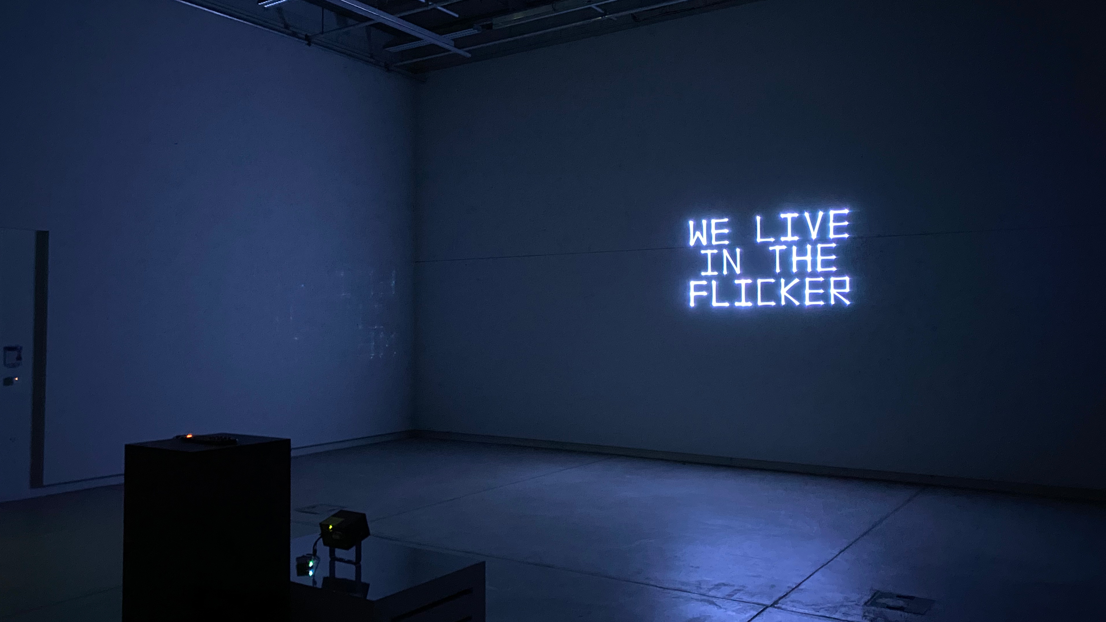 A laser projection onto a wall in a dark room. White text: We live in the flicker.