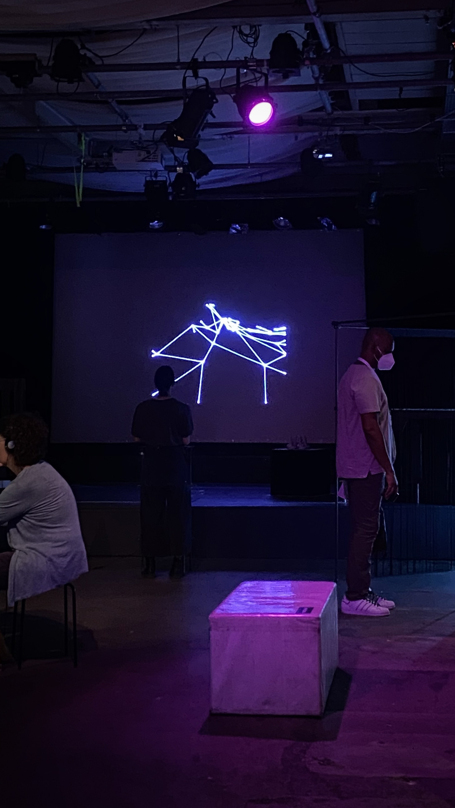 White abstract laser lines projected onto a screen on a stage. Purple lit room, people standing in front.