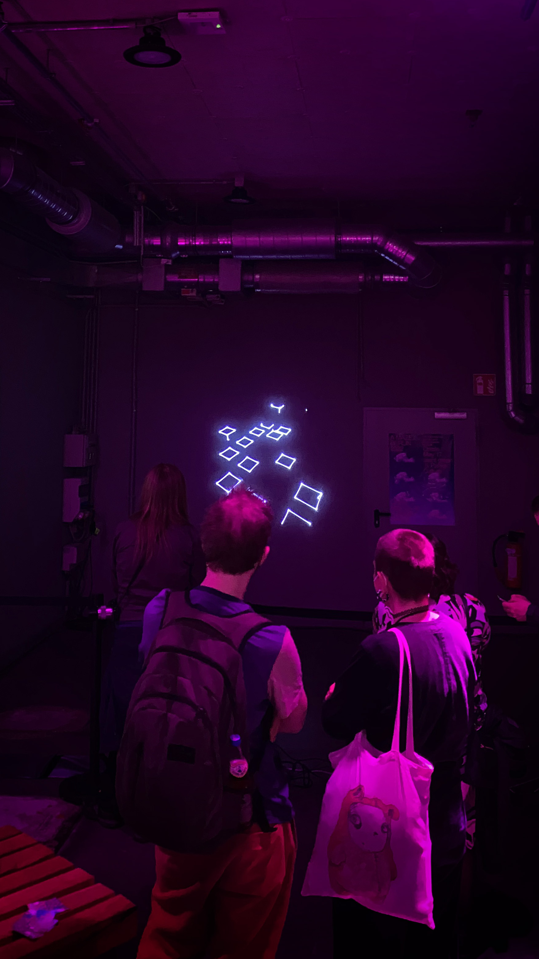 White laser light rhombi projected onto a wall. Purple lit room, people standing in front.